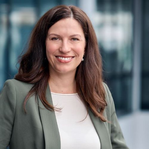 Porträt einer lächelnden Frau mit langen, braunen Haaren, in einem grauen Blazer vor einem unscharfen Hintergrund