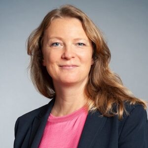 Porträt von Katja Fels mit blonden Haaren, lächelnd, in einem schwarzen Blazer und pinkem Oberteil, vor grauem Hintergrund.