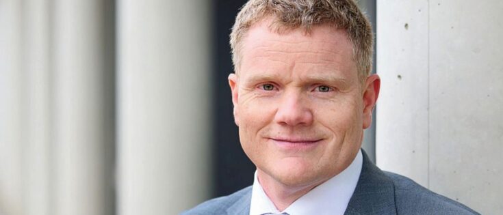 Mann in Anzug mit kurzen, blonden Haaren. steht vor einer hellen Wand und lächelt in die Kamera.