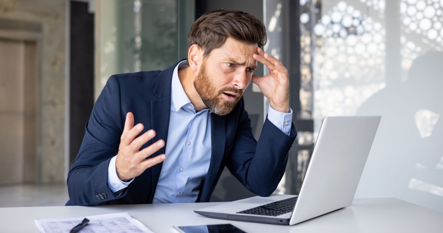 Junger Mann im Anzug sitzt am Schreibtisch, schaut verärgert auf den Laptop und hält sich den Kopf.