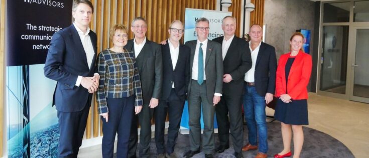 Gruppenfoto von acht Personen in einem modernen Büro, einige tragen Anzüge, andere legere Kleidung, mit H-Advisors-Logo ...