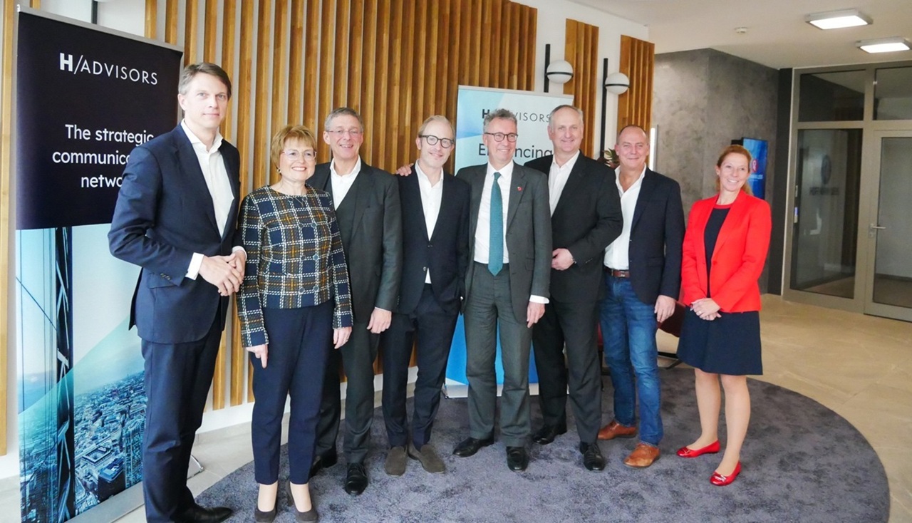 Gruppenfoto von acht Personen in einem modernen Büro, einige tragen Anzüge, andere legere Kleidung, mit H-Advisors-Logo ...