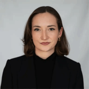 Porträt einer jungen Frau mit schulterlangen, braunen Haaren, in schwarzem Oberteil und Blazer, vor neutralem Hintergrund.