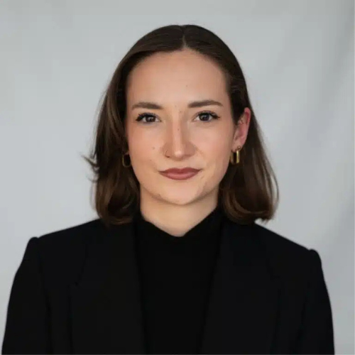 Porträt einer jungen Frau mit schulterlangen, braunen Haaren, in schwarzem Oberteil und Blazer, vor neutralem Hintergrund.