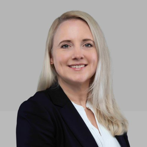 Porträt von Annika Hollmann mit schulterlangen, glatten blonden Haaren, lächelnd, in einem schwarzen Blazer vor grauem Hintergrund.