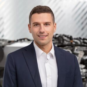 Junger Mann in Anzug lächelt vor unscharfem Hintergrund mit technischen Geräten.