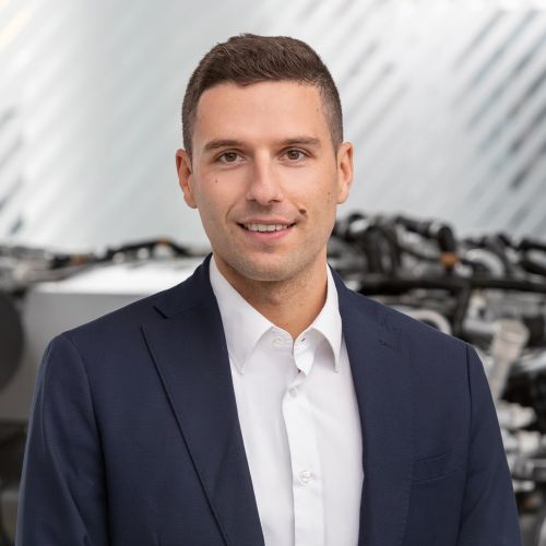 Junger Mann in Anzug lächelt vor unscharfem Hintergrund mit technischen Geräten.
