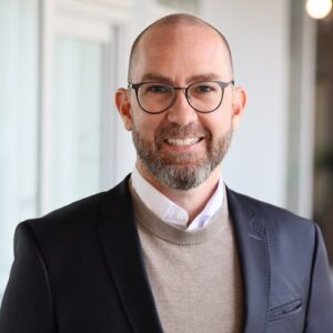 Mann mit Brille und Bart in einem Anzug lächelt vor einem modernen Hintergrund.