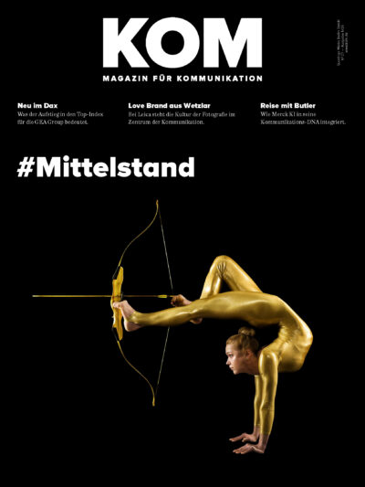 Magazin-Cover. Schwarzer Hintergrund. Davor eine Akrobatin in goldenem Ganzkörperanzug, die einen Handstand macht, mit ihren Füßen spannt sie einen Bogen, sie zielt. Titel "KOM - Magazin für Kommunikation" und Hashtag "#Mittelstand".