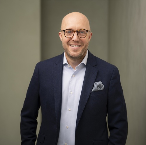 Mann mit Brille trägt einen dunklen Anzug und ein helles Hemd, lächelt vor neutralem Hintergrund.