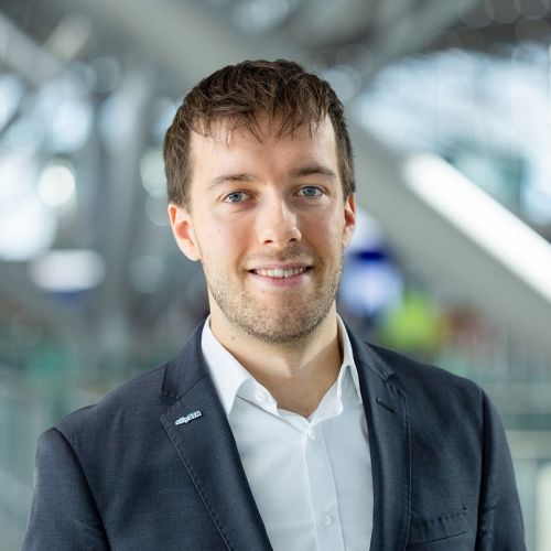 Mann mit kurzem Haar und Anzug lächelt vor moderner Flughafenarchitektur.