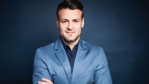 Mann in hellblauem Blazer mit verschränkten Armen vor einem dunklen Hintergrund.