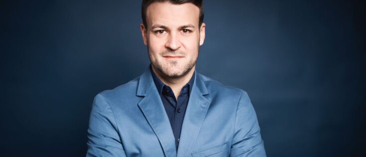 Mann in hellblauem Blazer mit verschränkten Armen vor einem dunklen Hintergrund.