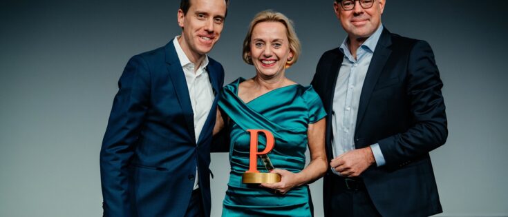 Drei Personen stehen auf einer Bühne, eine Frau in einem grünen Kleid hält einen Preis, die Männer tragen Anzüge.