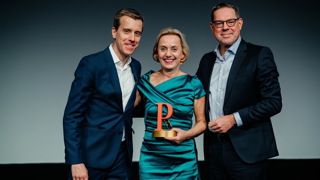 Drei Personen stehen auf einer Bühne, eine Frau in einem grünen Kleid hält einen Preis, die Männer tragen Anzüge.