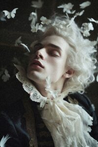 Porträt eines jungen Mannes mit blonden Locken, umgeben von weißen Schmetterlingen und Federn, in einem dramatischen Licht.