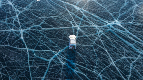 Ein Fahrzeug fährt über einen gefrorenen See mit sichtbaren Rissen im Eis und schneebedeckten Ufern im Hintergrund.