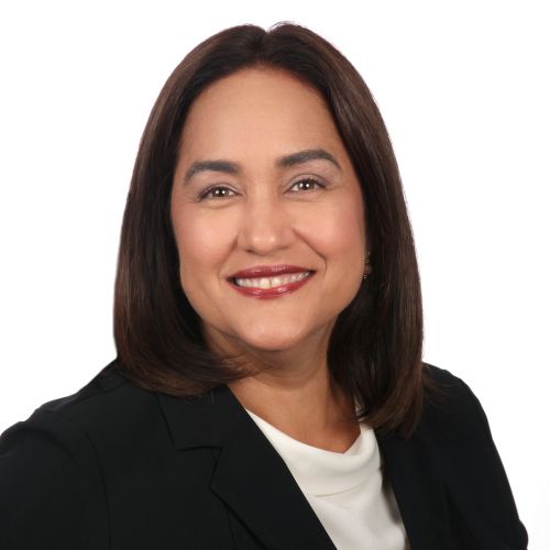 Porträt einer lächelnden Frau mit braunen Haaren, in schwarzer Jacke und weißem Oberteil, vor neutralem Hintergrund.