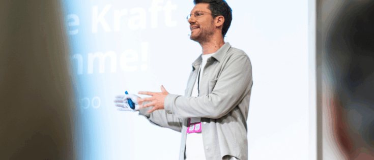 Mann spricht mit gestikulierenden Händen vor einem Bildschirm, auf dem der Text "Die Kraft der Stimme!" zu sehen ist.