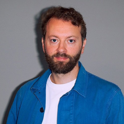 Mann mit Bart und lockigem Haar, trägt eine blaue Jacke und ein weißes T-Shirt, vor einem grauen Hintergrund.