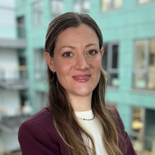 Frau mit langen, braunen Haaren, lächelnd vor einem modernen, hellen Hintergrund. Sie trägt einen burgunderfarbenen Blazer.