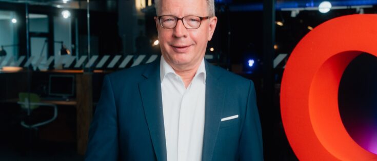 Mann in Anzug mit Brille steht vor einem großen roten Buchstaben "O" in einem modernen Raum.