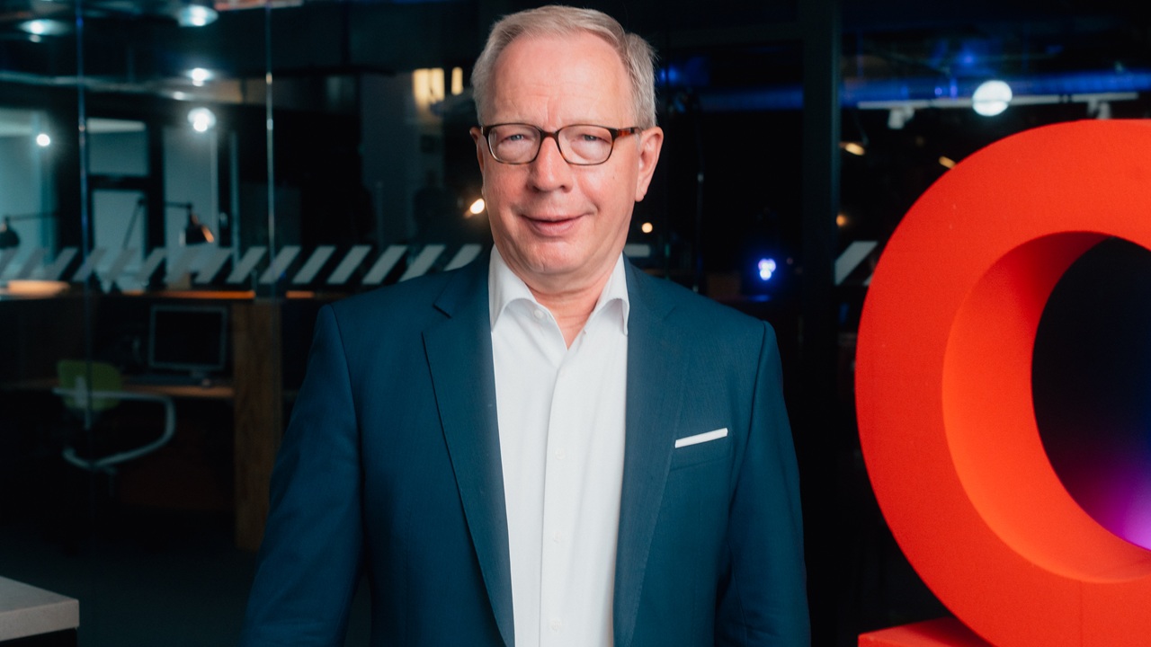 Mann in Anzug mit Brille steht vor einem großen roten Buchstaben "O" in einem modernen Raum.