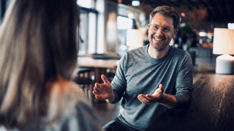 Mann mit Brille und grauem Pullover lächelt und gestikuliert während eines Gesprächs in einem modernen Café.