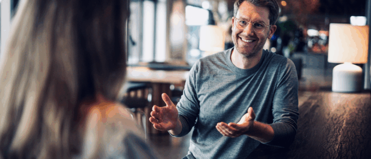 Mann mit Brille und grauem Pullover lächelt und gestikuliert während eines Gesprächs in einem modernen Café.