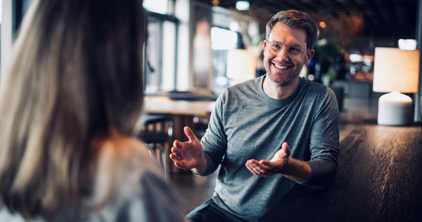 Mann mit Brille und grauem Pullover lächelt und gestikuliert während eines Gesprächs in einem modernen Café.