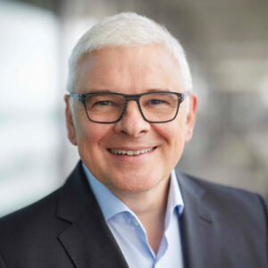 Porträt eines lächelnden Mannes mit grauen Haaren, Brille und Anzug, vor unscharfem Hintergrund.