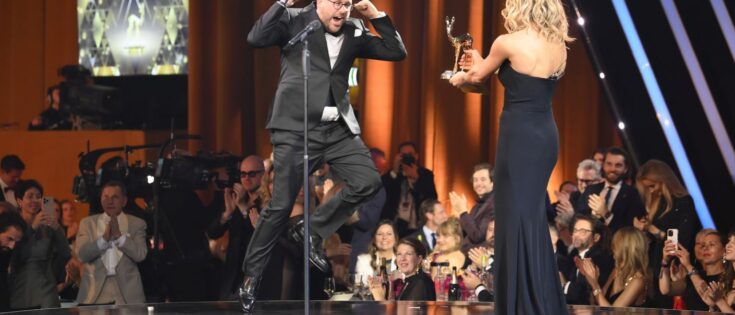 Paul Ronzheimer jubelt auf der Bühne, während Michelle Hunziker ihm einen BAMBI-Preis überreicht. Publikum applaudiert.