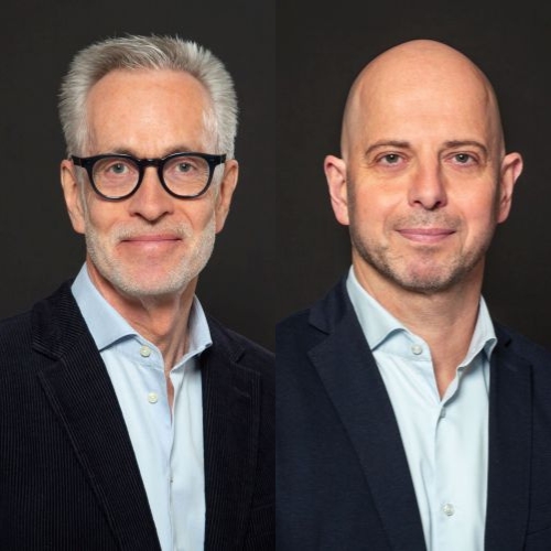 Zwei Männer in Anzügen vor einem dunklen Hintergrund, einer mit Brille und grauen Haaren, der andere mit Glatze.