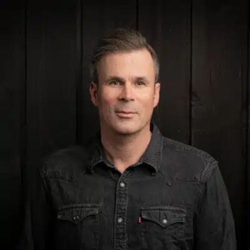 Porträt eines Mannes mit kurzen, grauen Haaren, in einem schwarzen Hemd vor einer dunklen Holzoberfläche.