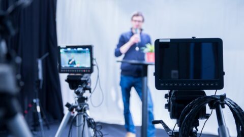 Männlicher Journalist spricht in ein Mikrofon, während Filmkameras im Vordergrund unscharf zu sehen sind.