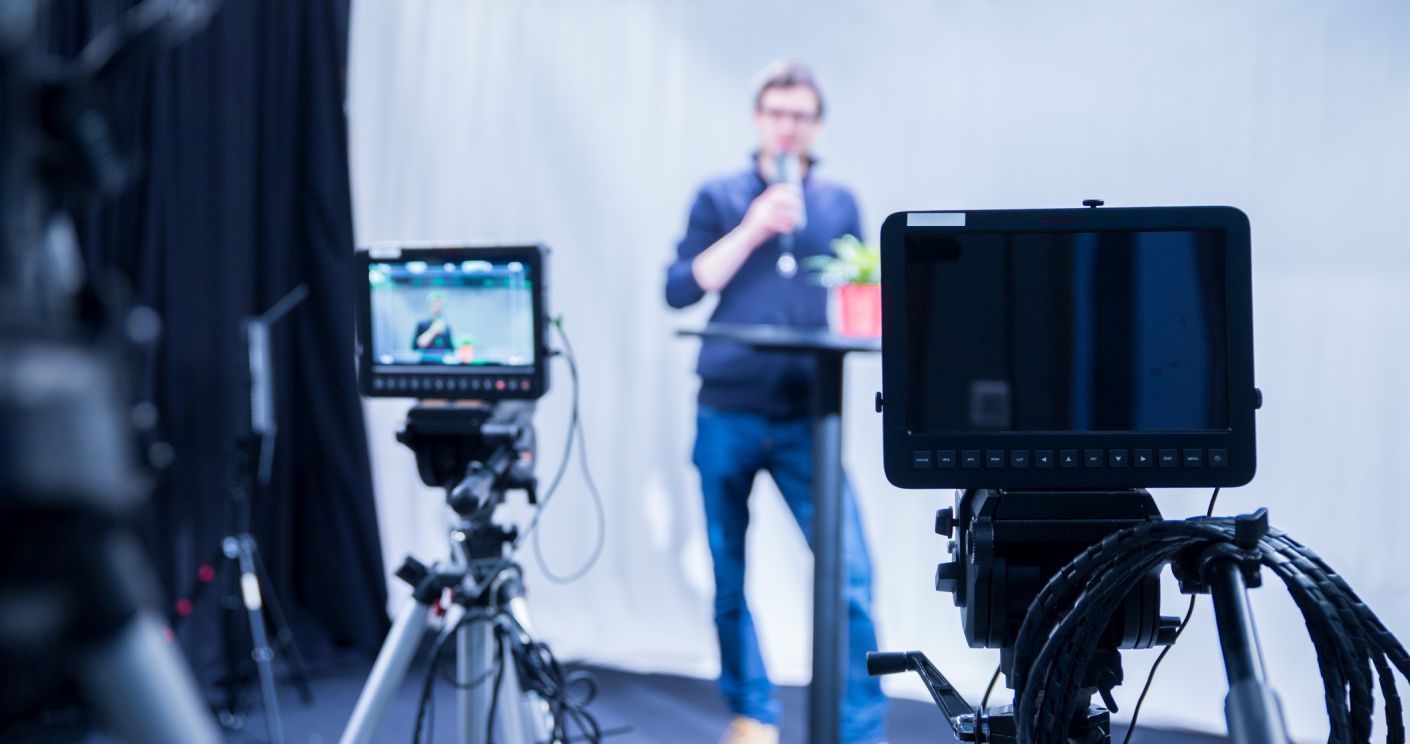 Männlicher Journalist spricht in ein Mikrofon, während Filmkameras im Vordergrund unscharf zu sehen sind.