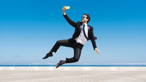 Geschäftsmann in Anzug springt akrobatisch und macht ein Selfie vor blauem Himmel und Meer.