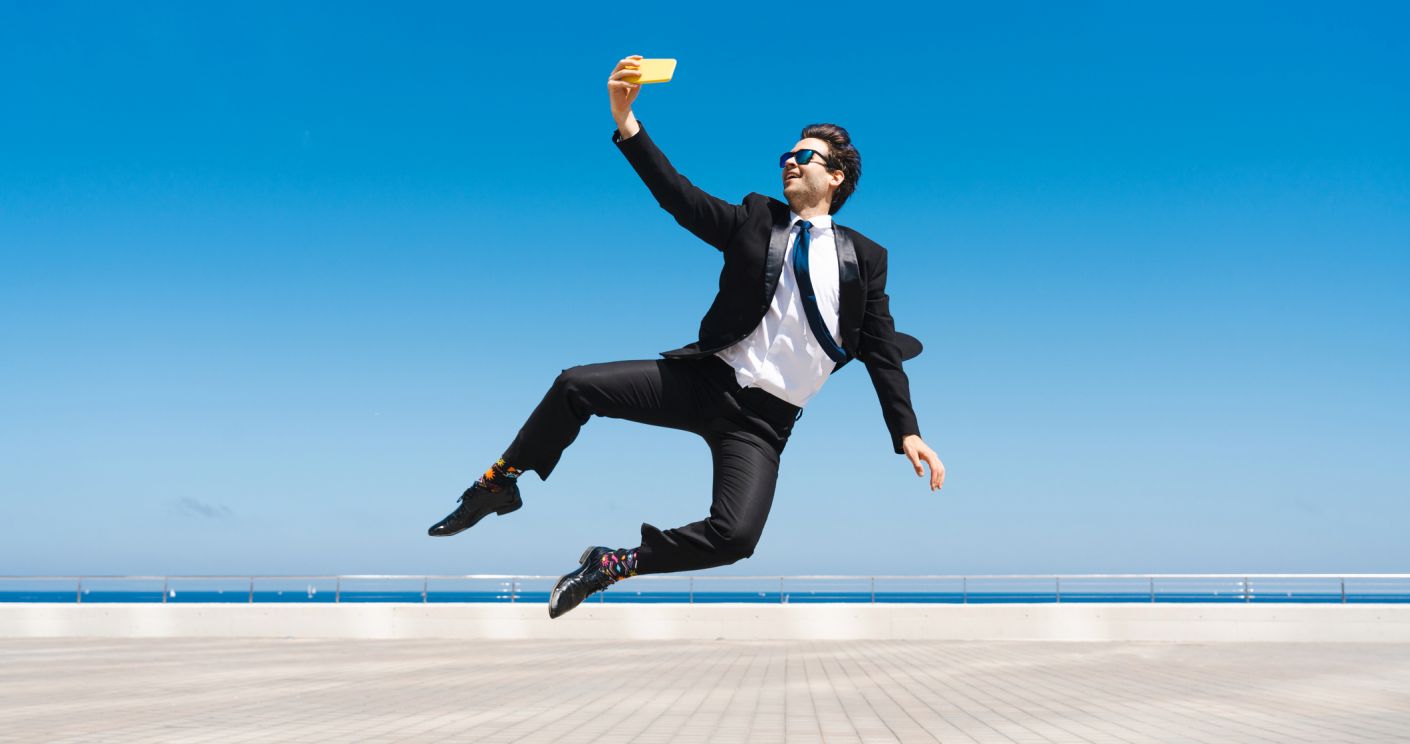 Geschäftsmann in Anzug springt akrobatisch und macht ein Selfie vor blauem Himmel und Meer.