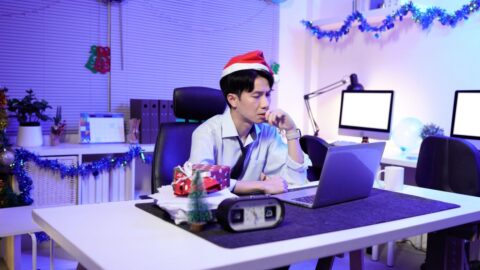 Geschäftsmann mit Weihnachtsmütze arbeitet spät im Büro, umgeben von Weihnachtsdekoration und einem Laptop.