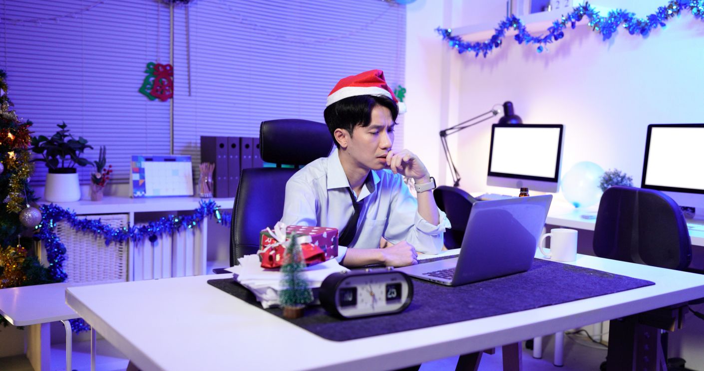Geschäftsmann mit Weihnachtsmütze arbeitet spät im Büro, umgeben von Weihnachtsdekoration und einem Laptop.