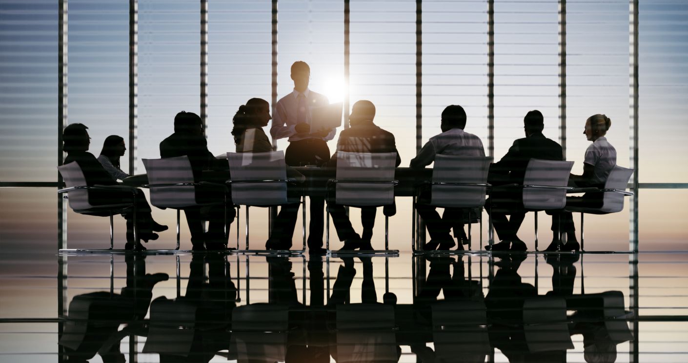 Silhouette von Geschäftsleuten in einem Konferenzraum, während ein Mann am Tisch spricht und Sonnenlicht durch ein bodentiefes Fenster scheint.