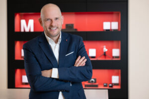 Mann in Anzug mit verschränkten Armen lächelt vor einem Regal mit roten Akzenten und Kameraausrüstung.