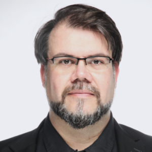 Porträt eines Mannes mit Brille, dunklem Haar und Bart, in schwarzer Kleidung vor hellem Hintergrund.