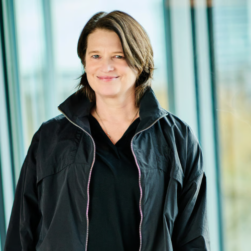 Porträt von Kerstin Neuber in schwarzer Jacke, lächelnd vor einer modernen Glasarchitektur.