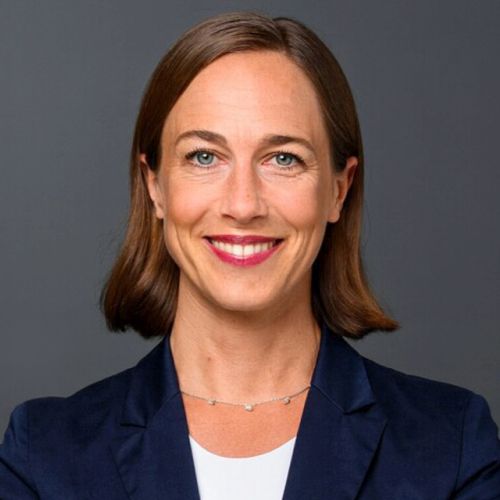 Porträt von Carolin Strunz mit kurzem, braunem Haar, blauer Blazer und freundlichem Lächeln vor grauem Hintergrund.