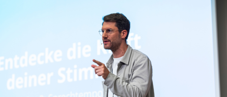 Mann mit Brille spricht auf einer Bühne, im Hintergrund ein Bildschirm mit dem Text „Entdecke die Kraft deiner Stimme“.