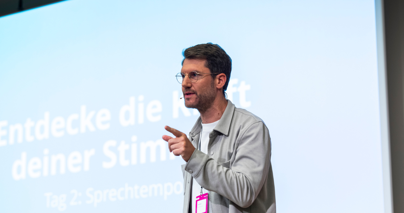 Mann mit Brille spricht auf einer Bühne, im Hintergrund ein Bildschirm mit dem Text „Entdecke die Kraft deiner Stimme“.