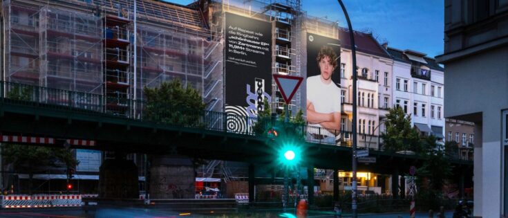 Stadtansicht mit Bauarbeiten, Plakat eines jungen Mannes an einer Brücke, Verkehrssignal und Autos im Vordergrund.
