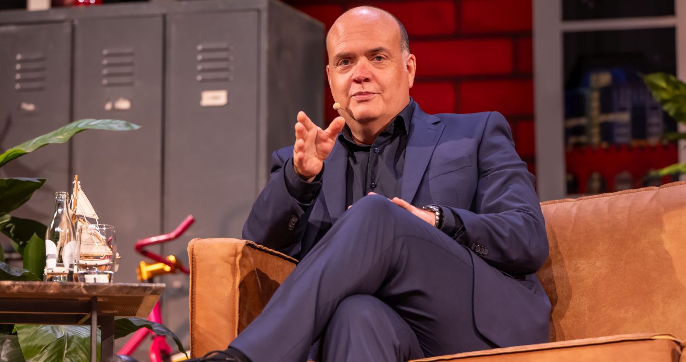 Mann in Anzug sitzt auf Sofa, spricht mit gestikulierender Hand, im Hintergrund Pflanzen und Regale.