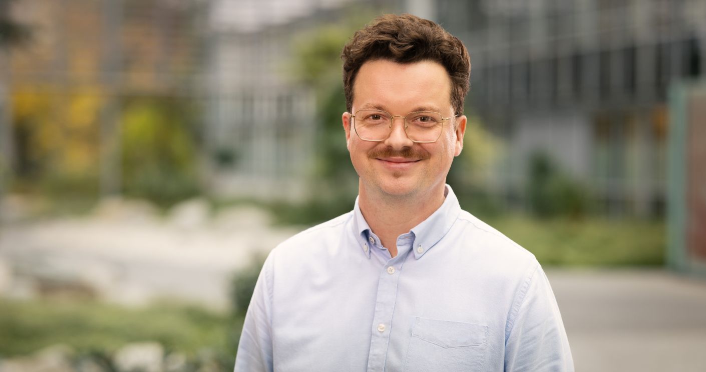 Lächelnder Mann mit Brille und Schnurrbart, trägt ein weiß-bläuliches Hemd, steht vor einem modernen Hintergrund.